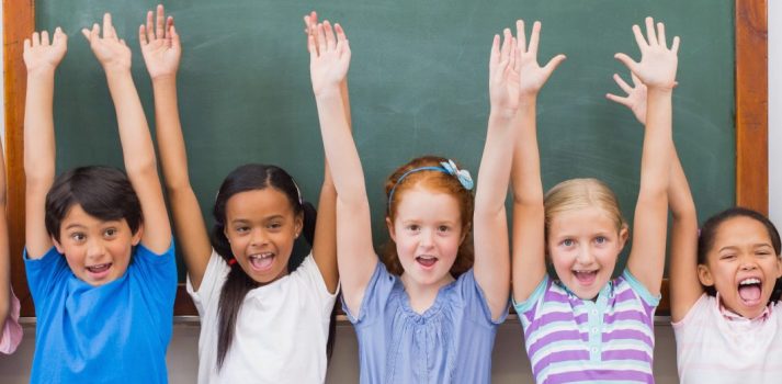 acht kleine Kinder stehen vor Tafel und halten ihre Arme in die Luft