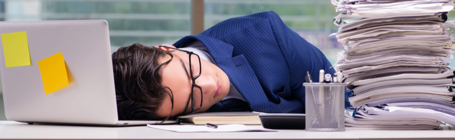 Mann im Anzug schläft am Arbeitsplatz neben einem großem Papierstapel